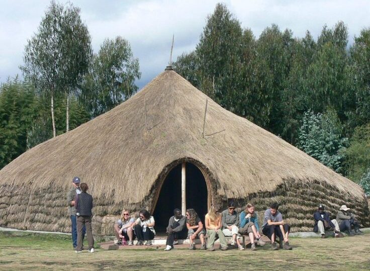 A large, traditional Rwandan thatched-roof structure with a group of tourists seated outside at the Gorilla Guardians Village in Kinigi by excellent african safaris.